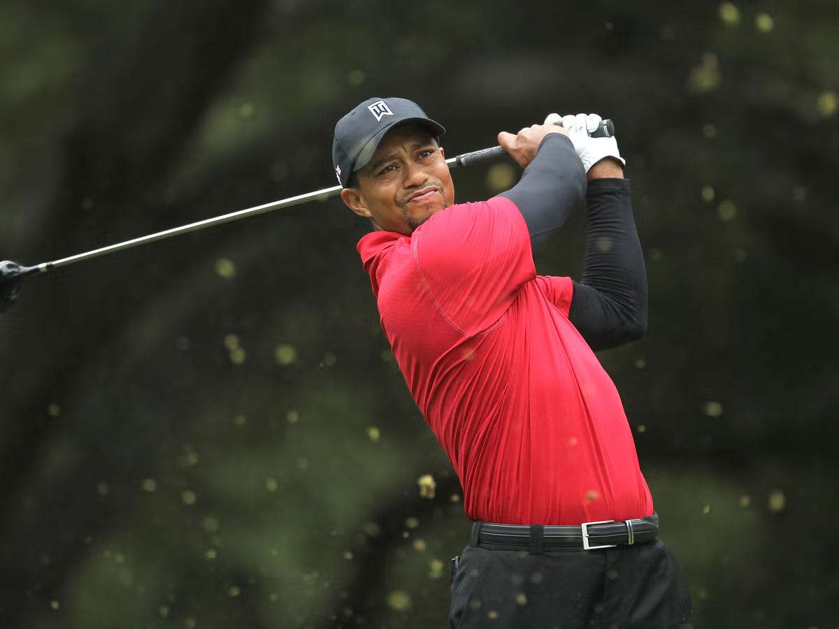 Tiger Woods mid-swing on a golf course, wearing a red shirt and black cap, with trees in the background.