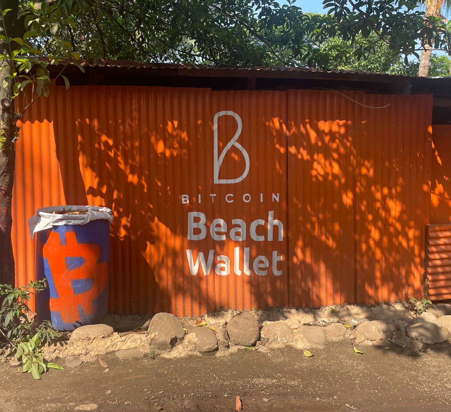 White writing on an orange corrugated metal building reads "Bitcoin Beach Wallet." The Bitcoin logo has been spray painted onto a trash can beside it.