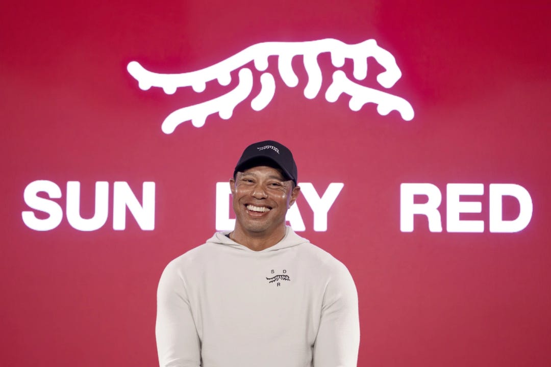 Tiger Woods smiling in a white hoodie and black cap, standing in front of a red background with the "Sun Day Red" logo and text.