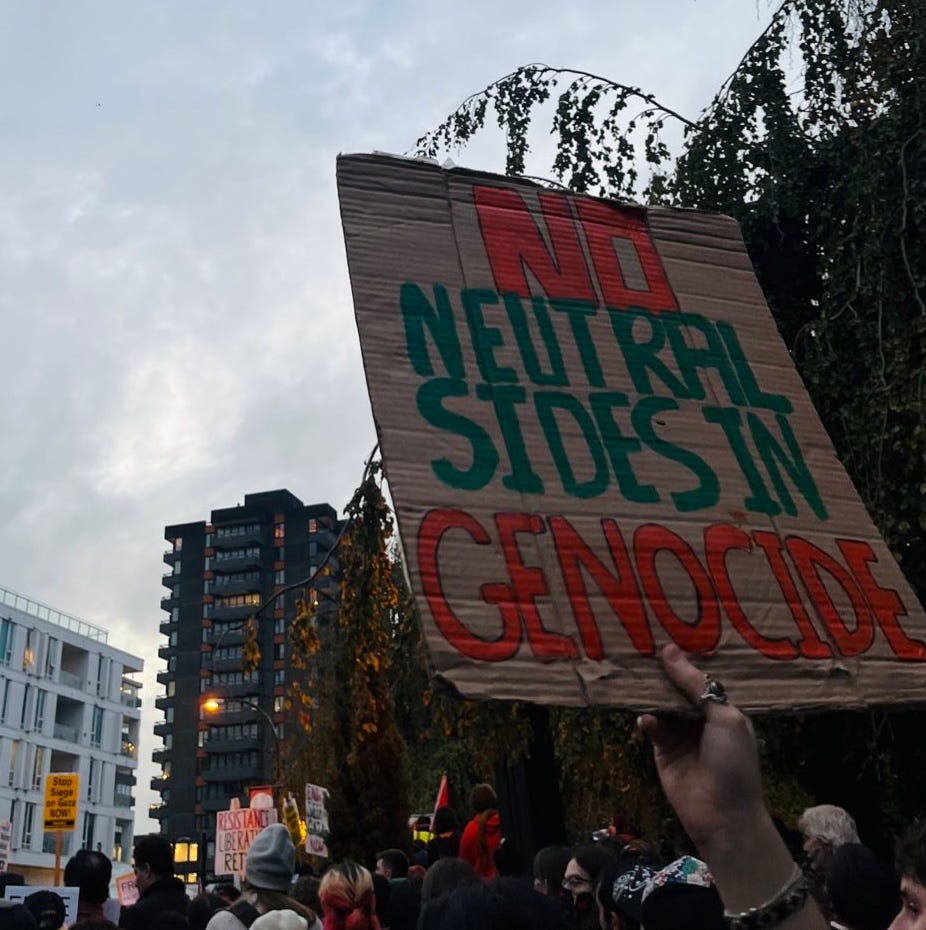 At an anti-genocide protest, a protester holds a placard reading "No neutral sides in genocide" in red and green lettering