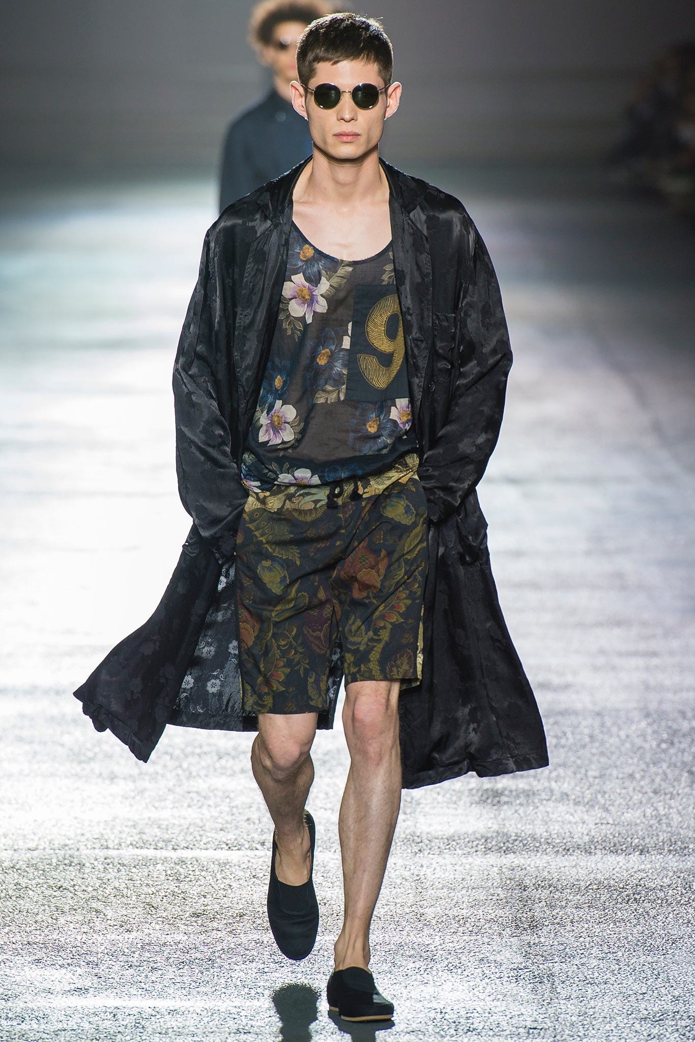 A model on a fashion catwalk, who is a white man with dark brown hair, wearing a loosely fitted floral t-shirt with a low scoped neckline, dark floral shorts, and an unbuttoned knee length raincoat, with slip-on plimsolls. A model on a fashion catwalk, who is a white man with dark brown hair, wearing a loosely fitted floral t-shirt with a low scoped neckline, dark floral shorts, and an unbuttoned knee length raincoat, with slip-on plimsolls.