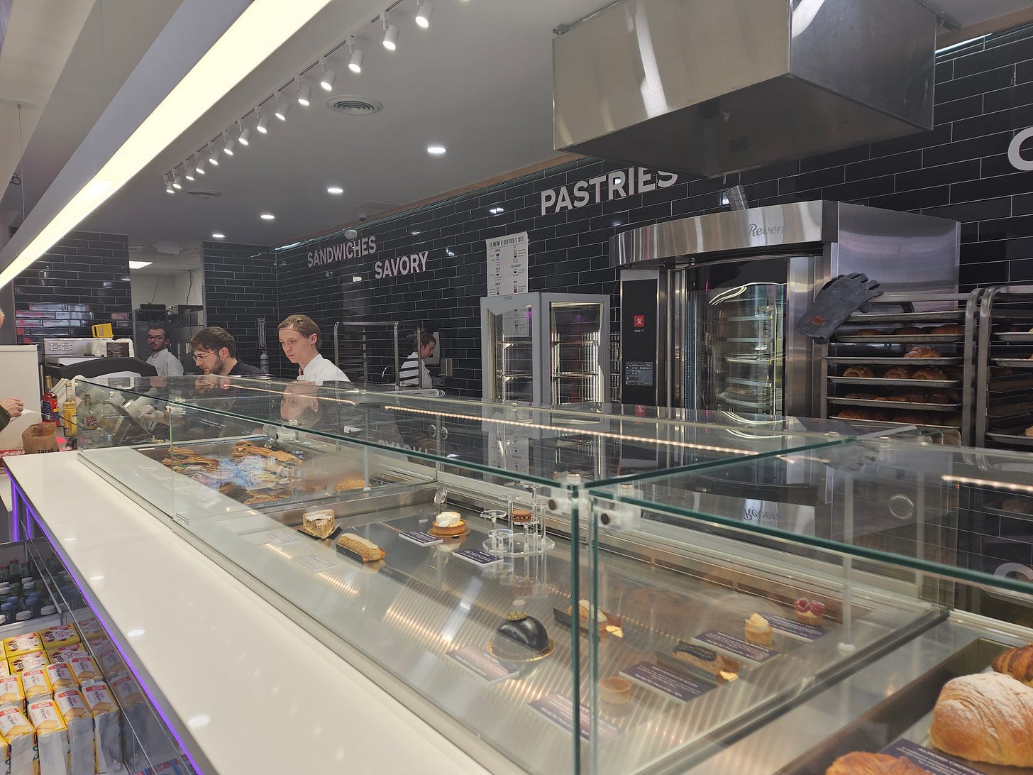 Picture shows the glass gallery with pastries in Fresh Baguette. There is ceiling lighting that is illuminating the area behind the counter with servers. A coffee machine can also be seen and a customer who is ordering.