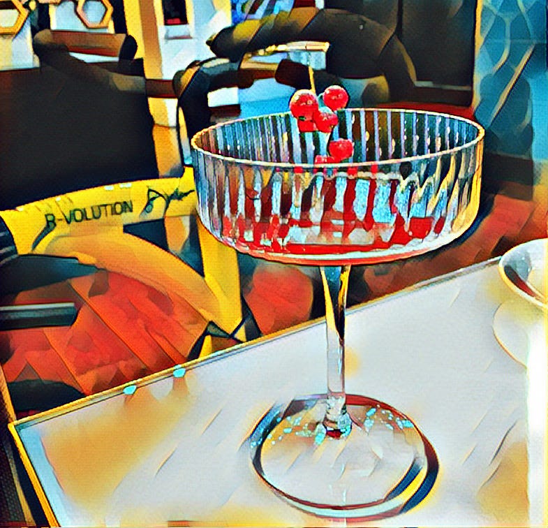 Foreground is an almost finished cocktail on a restaurant table - pinky red liquid in an angular champagne saucer style glass, with a sprig of cranberries in it. Behind it is Jane’s Alinker, a bright yellow walking bike with black trim. Image is stylised to give a pop art feel. 