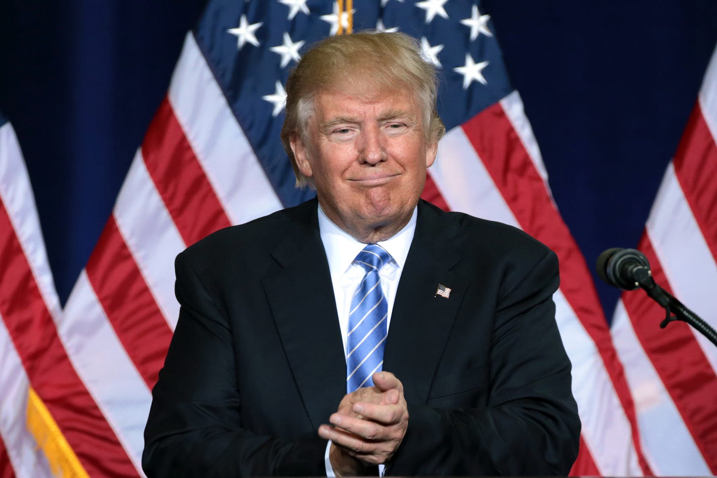 President Trump wearing a black suit and blue tie standing in front of a row of American flags