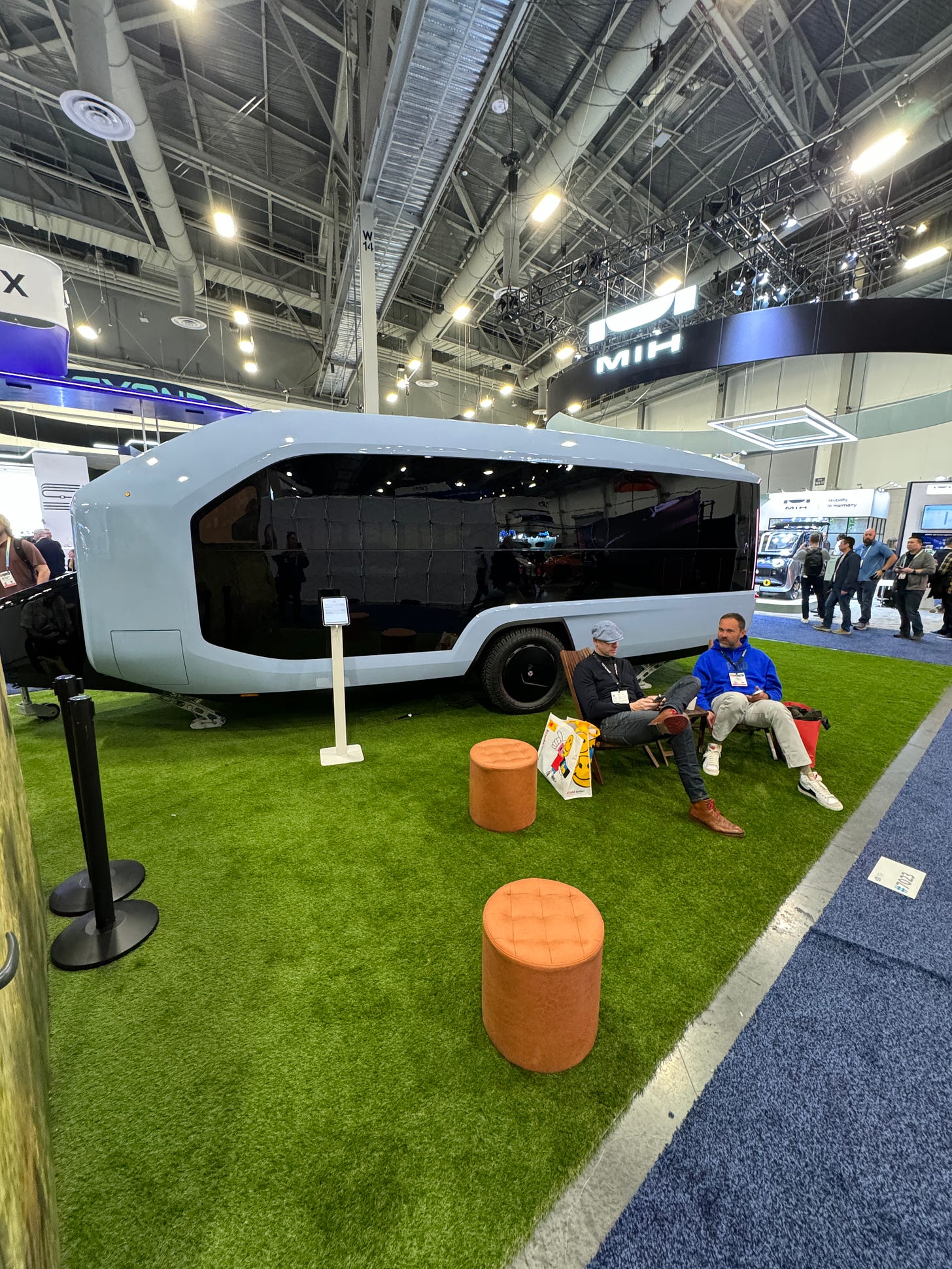 A sleek blue trailer with black windows that wrap all the way around, parked in a convention center on faux grass with conference-goers hanging out on chairs in front of it.