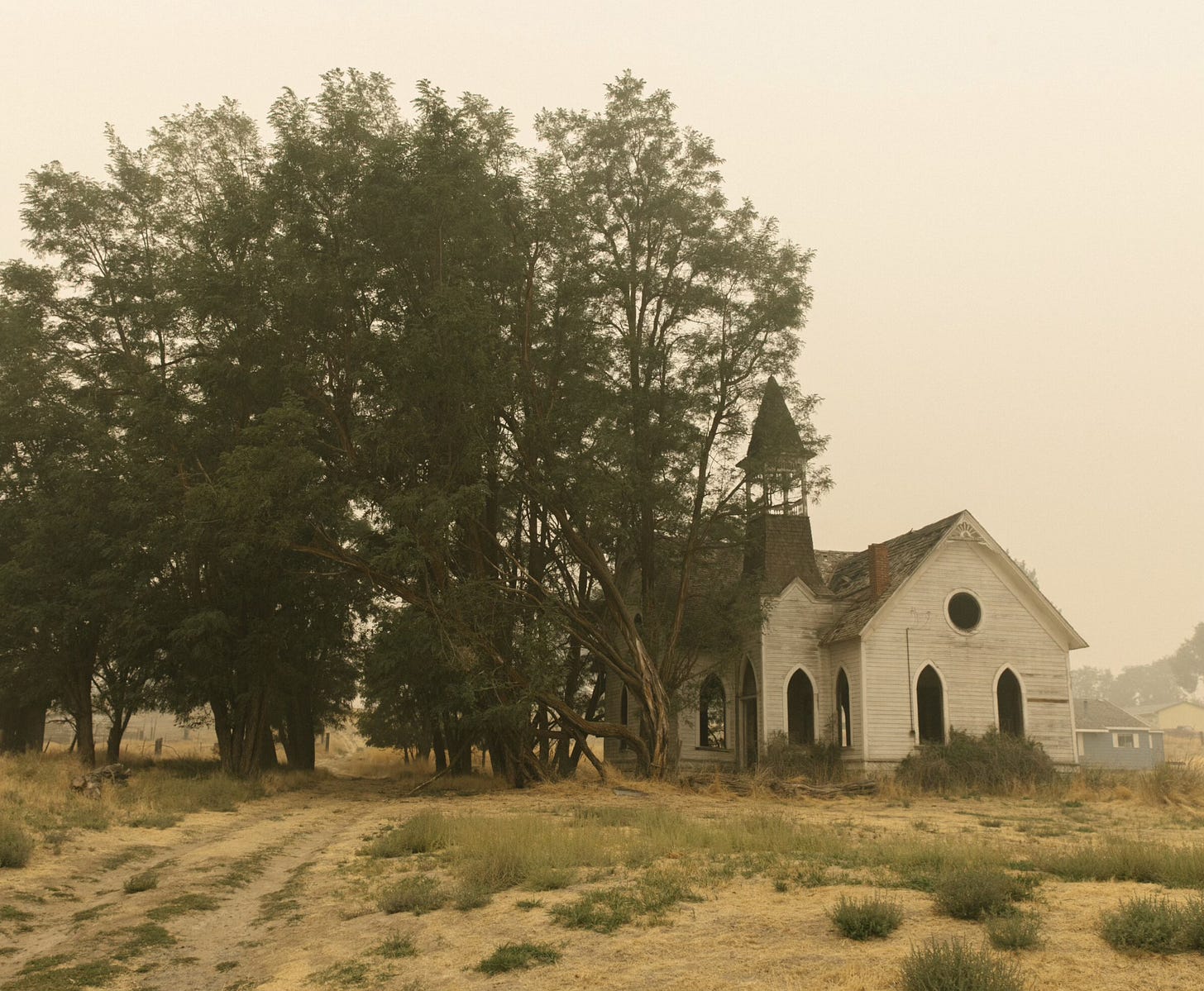 An old, weathered church with Gothic-style arched windows sits beside large trees on a dirt path. The sky is hazy, and the grass is dry.