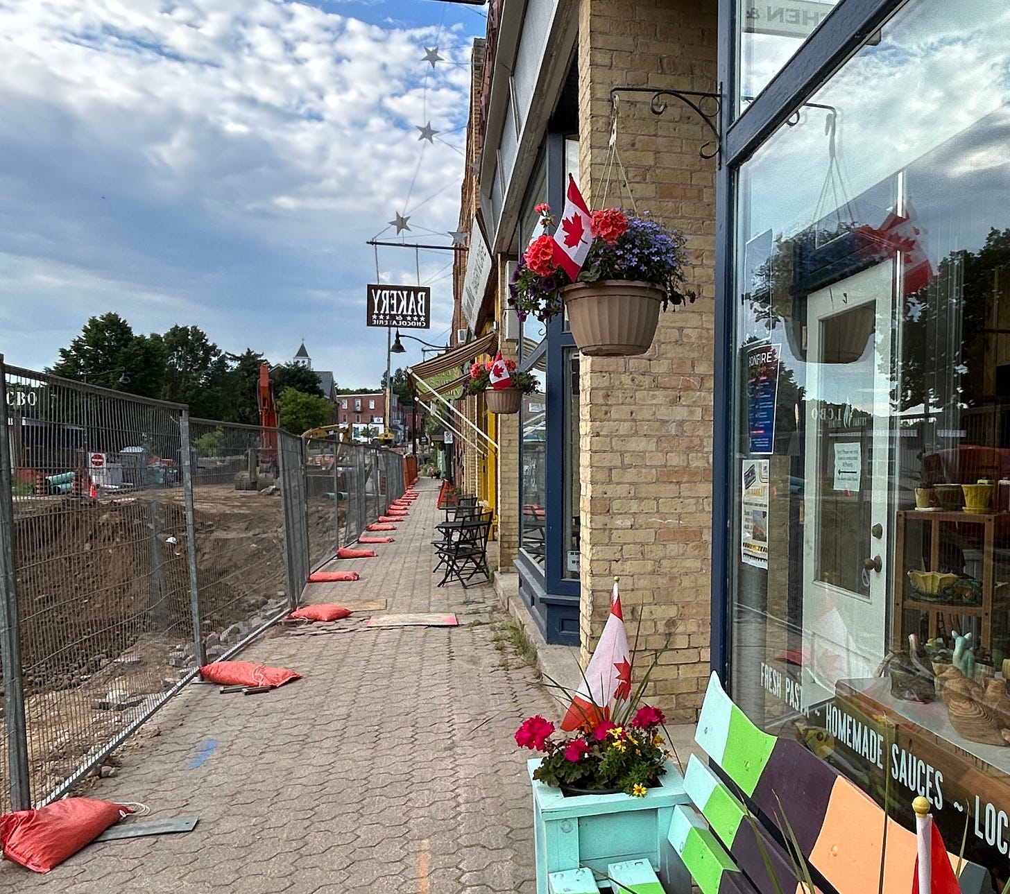 A street under construction with fencing between the road and sidewalk and businesses along the right side.