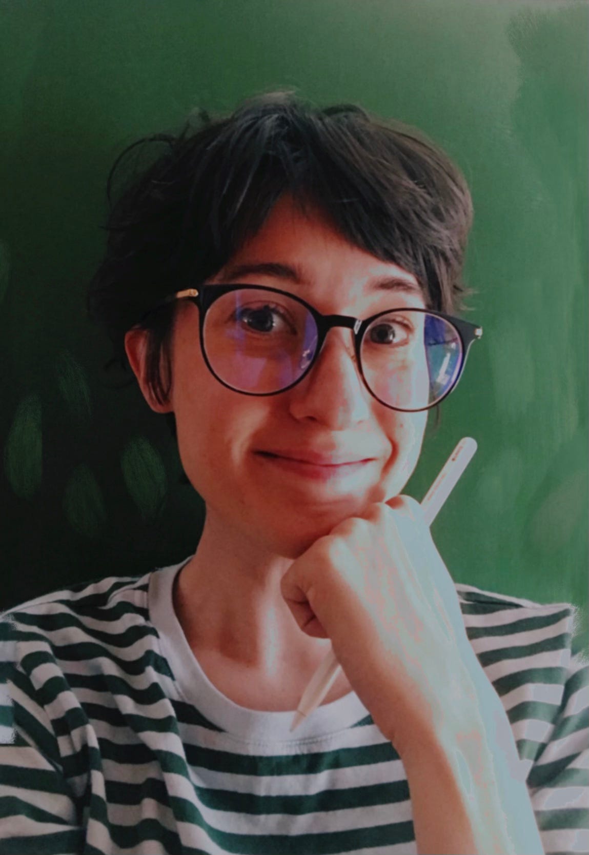 Gracie, a brown-haired person with black glasses and a striped shirt, smiles at the camera, holding a pencil and supporting her chin with the their hand