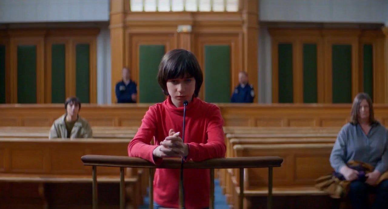 Movie still from Anatomy of a Fall. A young boy stands to speak in a nearly empty courtroom.