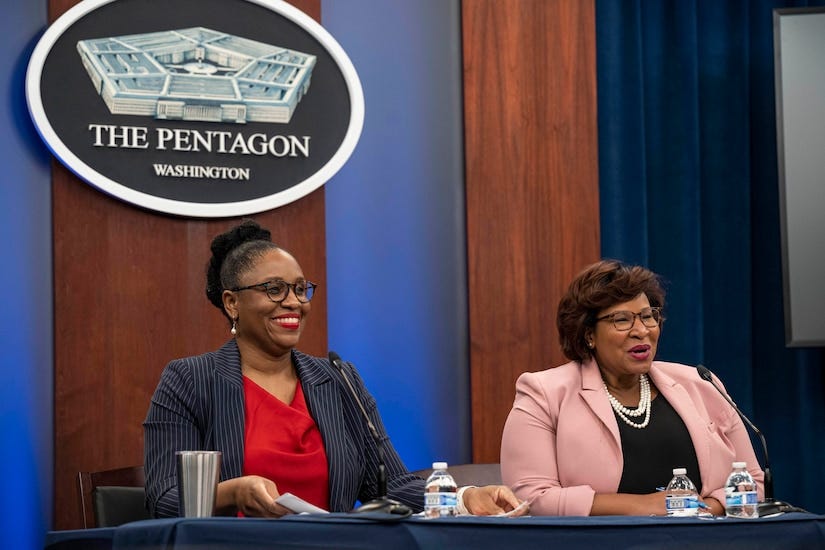 Two smiling women wearing business attire sit at a table with the Pentagon seal behind them.