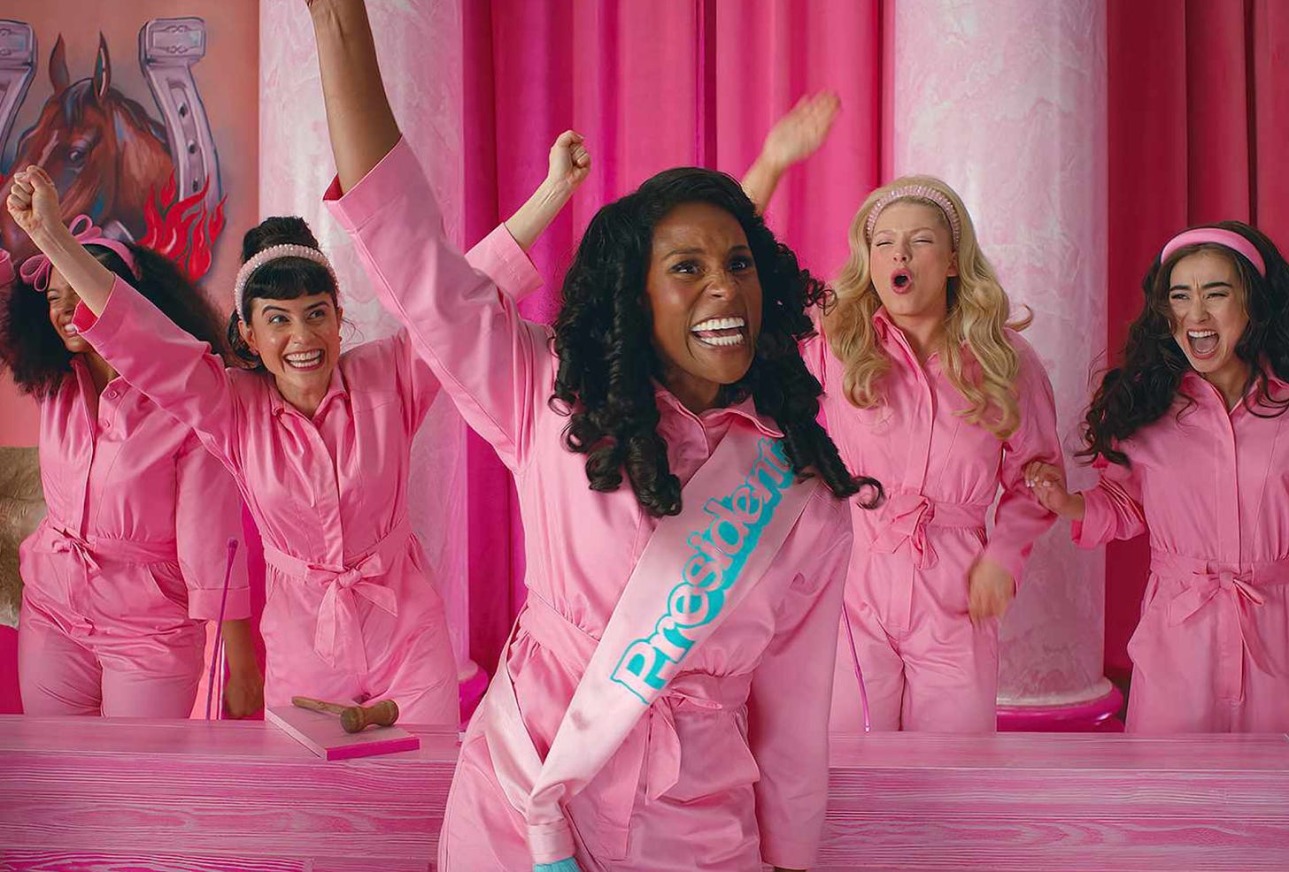 Issa Rae as President Barbie, wearing a pink jumpsuit and sash, raising her arm and cheering while other Barbies cheer behind her