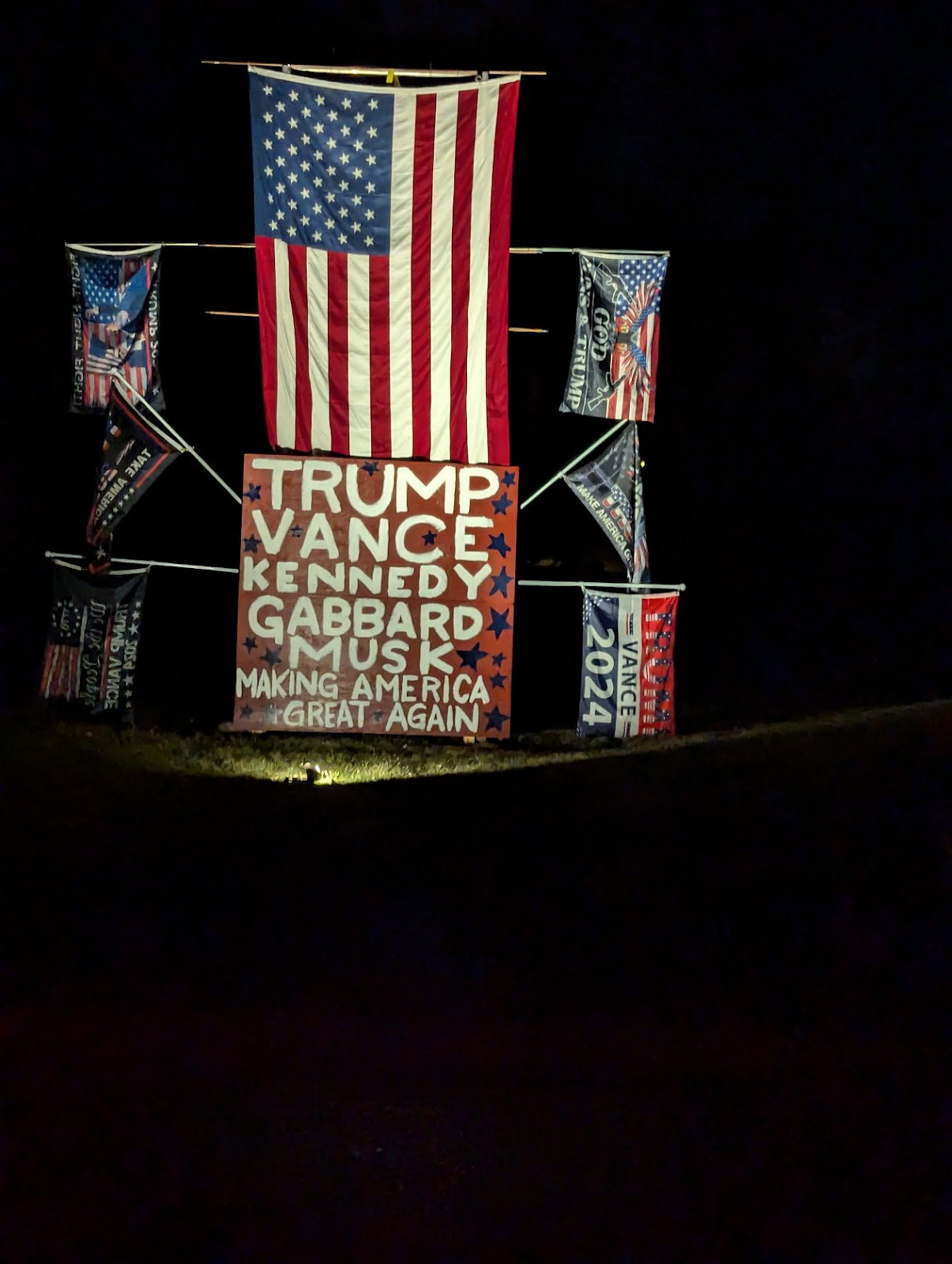 A series of MAGA flags arranged in an altar-like configuration. At the center, lit by a spotlight, is spray painted: Trump Vance Kennedy Gabbard Musk Making America Great Again