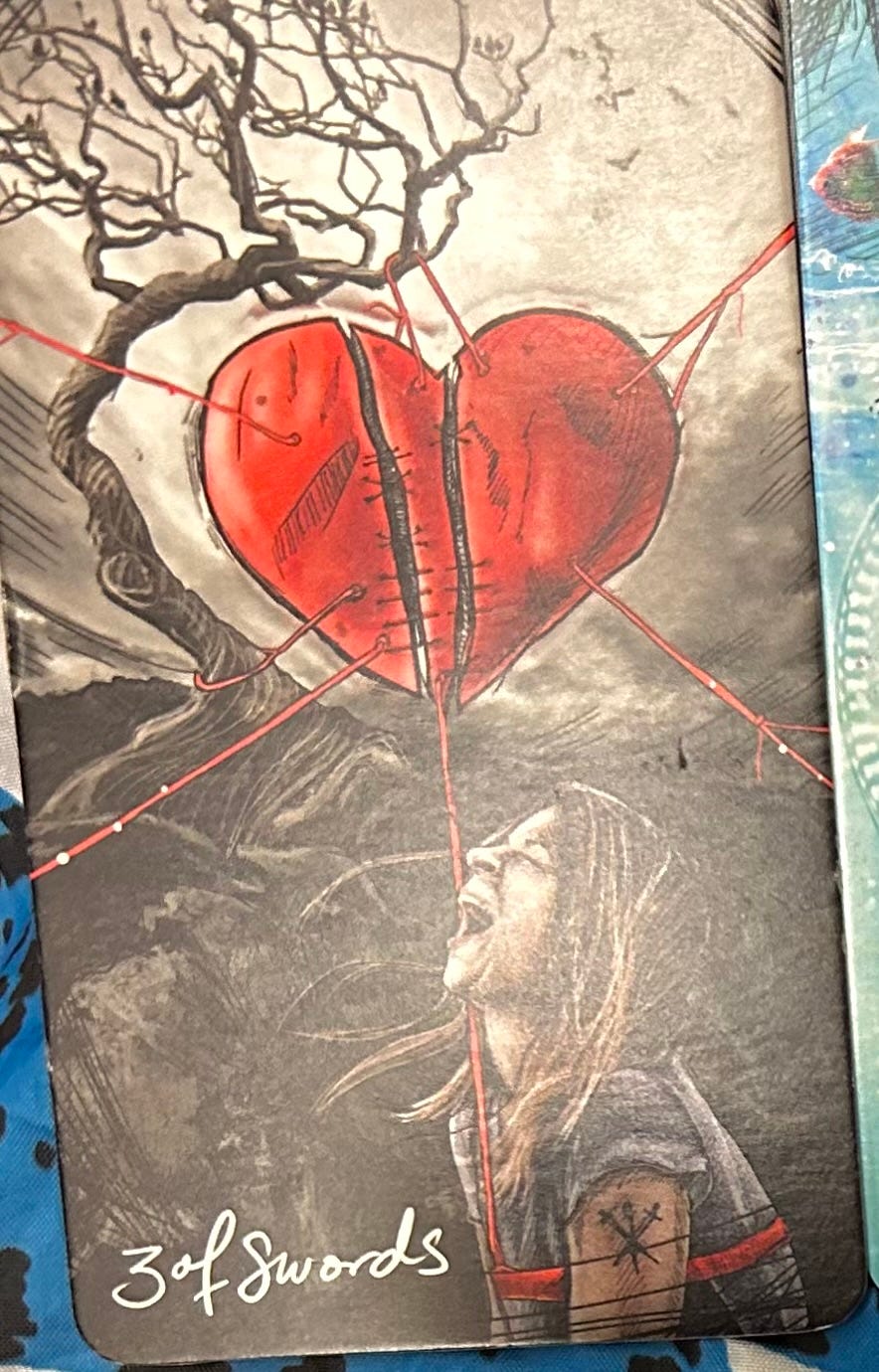 A large, red, stitched-together, broken heart hangs from a bare tree with a screaming person with long hair sitting underneath.