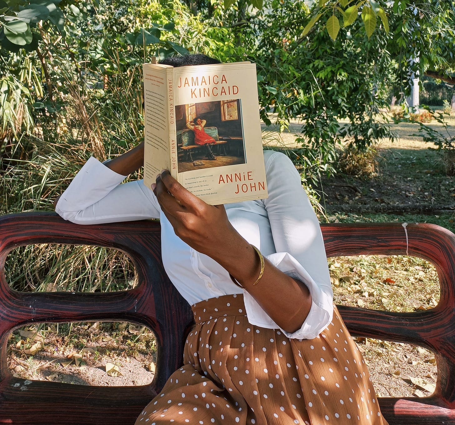 A black woman in a brown and white polka dotted skirt and long sleeve white shirt holding a paperback copy of Annie John in front of her face as she sits on a red bench under the tree. The book cover is pale brown with a detail from a painting of a black girl sitting on a bench in a room by a window.