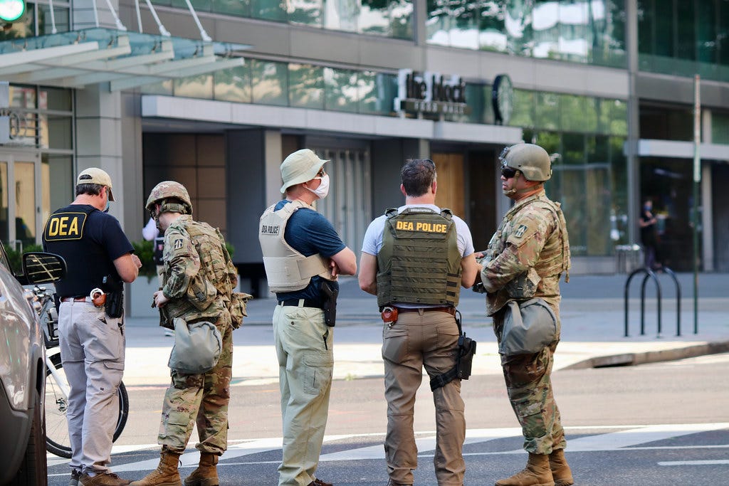 Several armed persons in D.E.A. vests or camouflage standing in a street in front of a building 