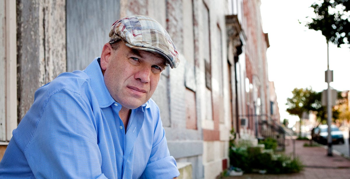 This image shows a photo of author, journalist, & television scribe David Simon wearing a newsboy cap and sitting on the stoop of a Baltimore, Maryland rowhouse in 2010. This image shows a photo of author, journalist, & television scribe David Simon wearing a newsboy cap and sitting on the stoop of a Baltimore, Maryland rowhouse in 2010.