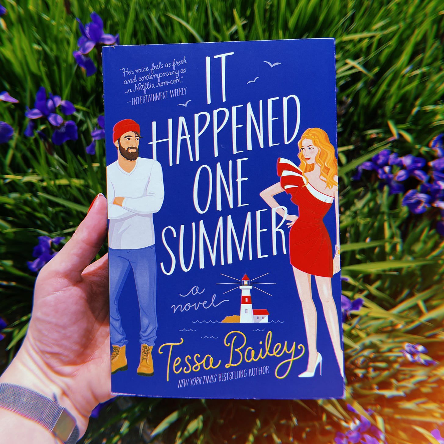 A white woman's hand holding a book, It Happened One Summer by Tessa Bailey, in front of some purple flowers.