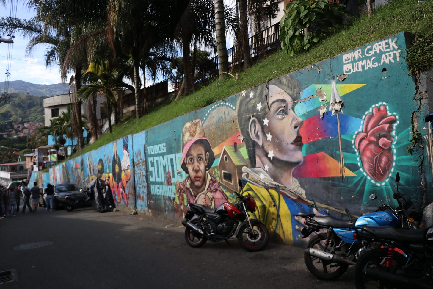 Motorcycles stand parked before a wall covered in street art showing a heart, several faces, and the words "todos somos migrantes."