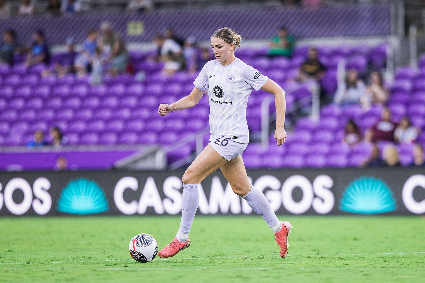 Women with brown hair in a pony tail in profile and a purple and white kit dribbles a soccer ball