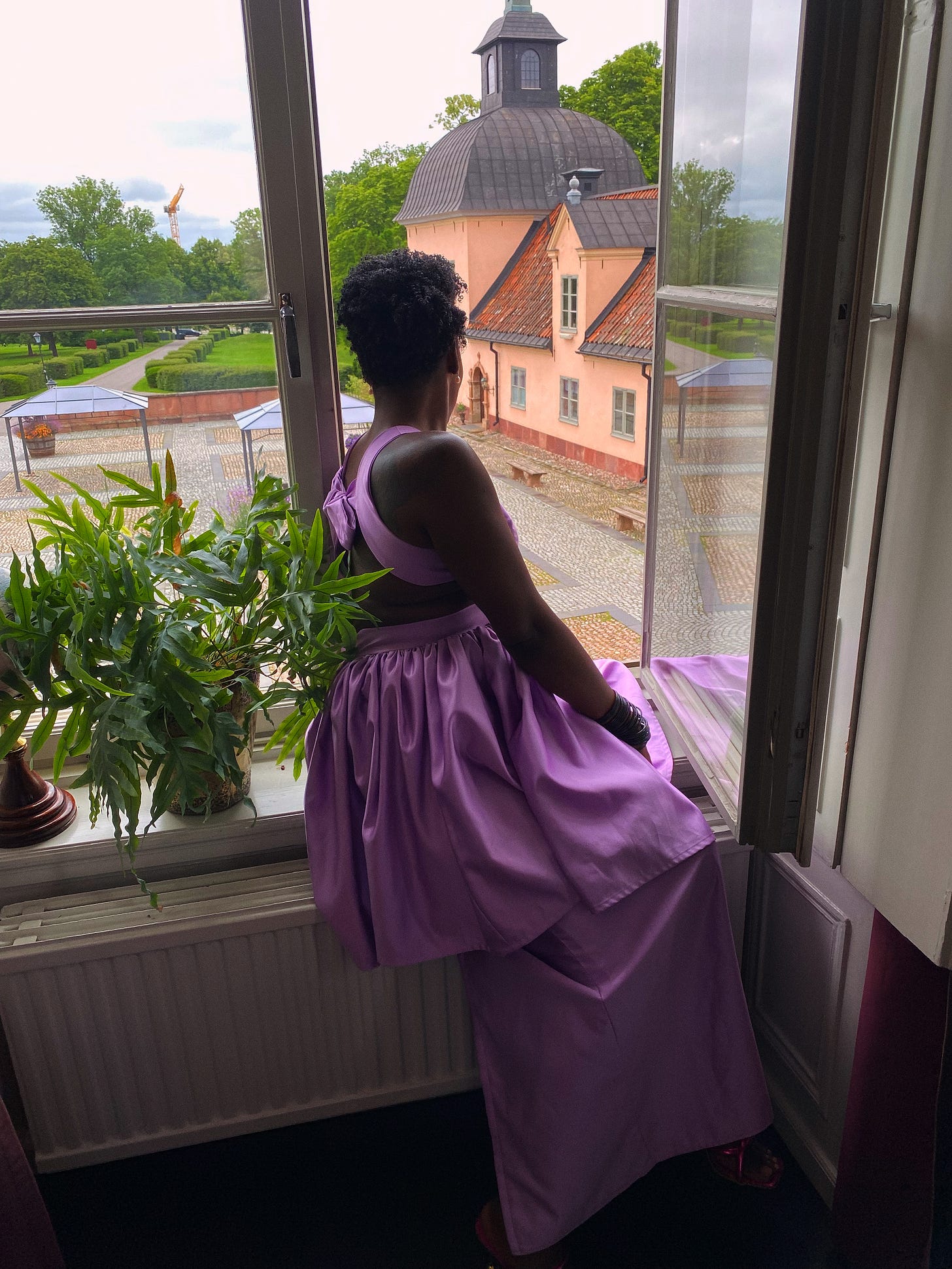 Lovette Jallow seated, shot from a low angle, holding a glass and wearing lavender. Lovette Jallow seated, shot from a low angle, holding a glass and wearing lavender.