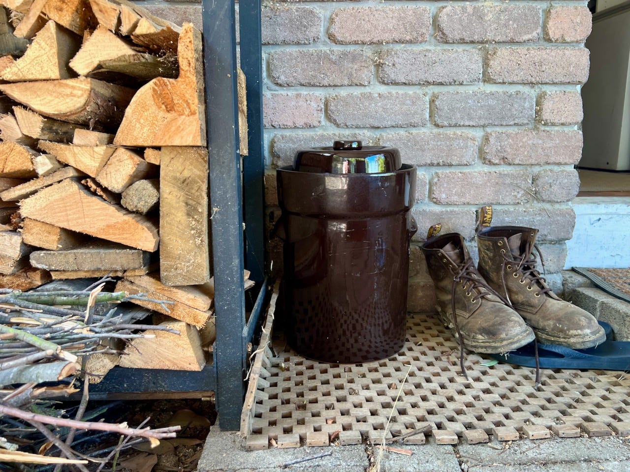 A browen fermentation crock infront of a brick wall by a door, with firewood and boots beside it A browen fermentation crock infront of a brick wall by a door, with firewood and boots beside it