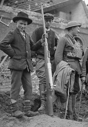 Black and white photo of British officer with civilians in Bohain, 10 October 1918. A boy of may 10 years old is holding a large anti-tank rifle and looking very pleased with himself.