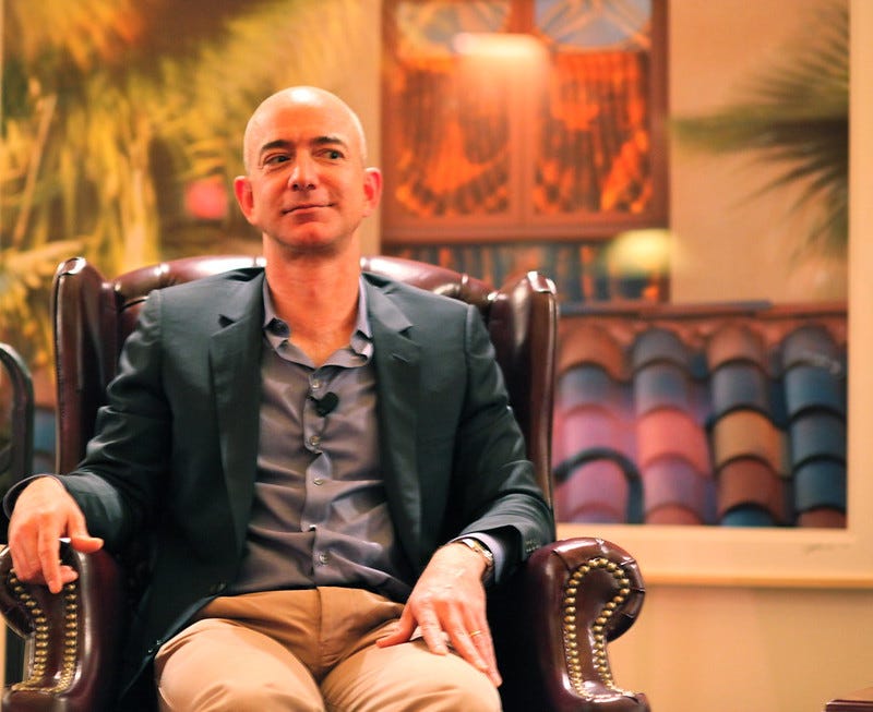 Photo of Amazon's Jeff Bezos seated in a leather chair looking side-eyed at some person off-carema. Bezos is dressed casually in a jacket and gray shirt with no tie, wearing a lapel microphone clipped to the shirt.