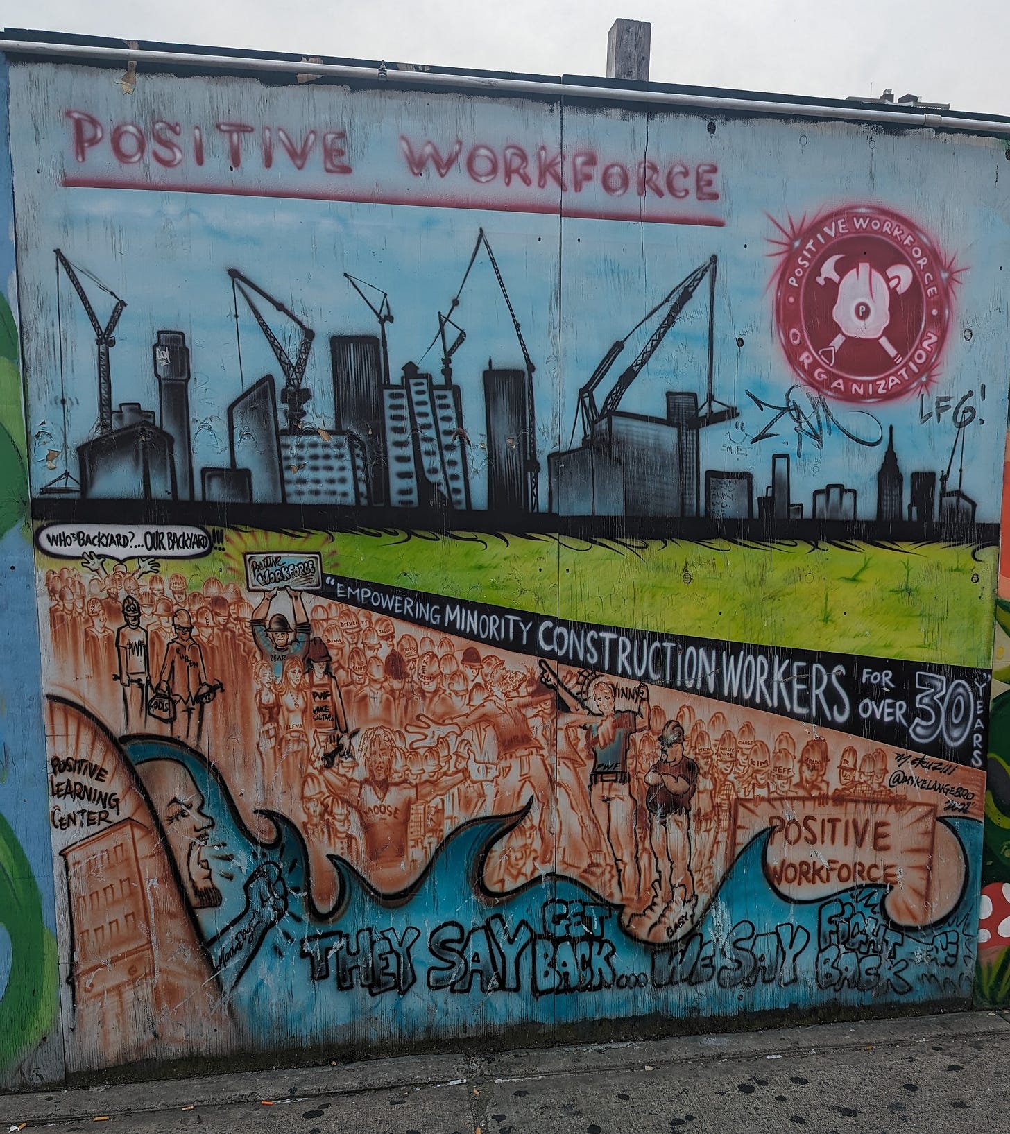A painted wood fence that reads: Positive Workforce. Empowering Minority Construction Workers for over 30 years. They say Get Back. We say Fight Back.