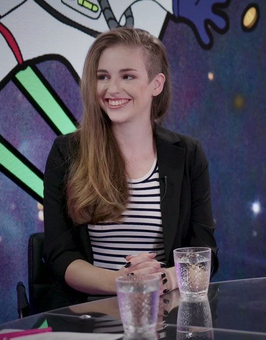 A photograph of Molly White sitting at a table in a video studio. She's       a white woman with long brown hair that's shaved on one side, she has brown eyes, and she's wearing a black and       white striped shirt and black blazer. There is a colorful cartoon background behind her. She's smiling and looking       at someone out of frame.