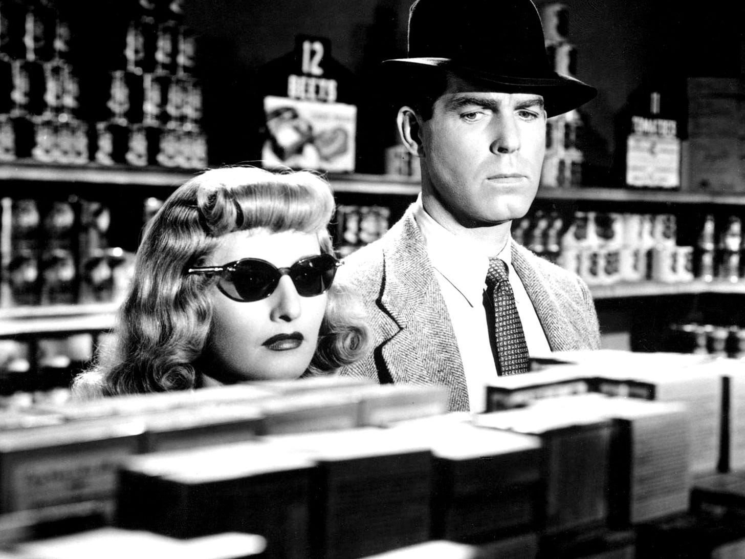 Barbara Stanwyck and Fred MacMurray in a supermarket in 1944 film noir Double Indemnity. Stanwyck is wearing dark glasses and we only see her head above the supermarket shelving. MacMurray is wearing a black fedora, a soft tweed blazer and a check tie. They both look suspicious. They are plotting a murder