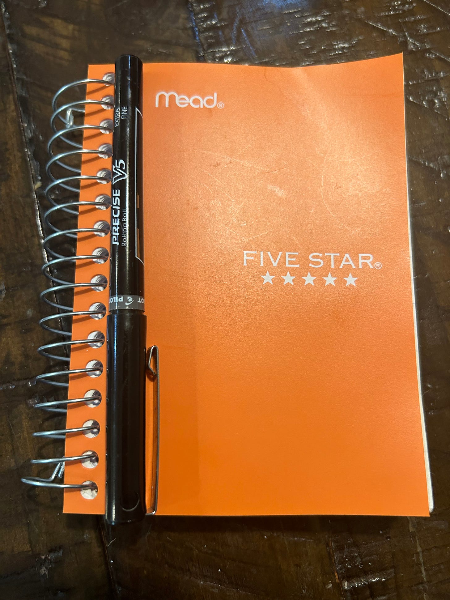 An orange spiral notebook resting on a brown table, with a black pen resting alongside the spirals. 