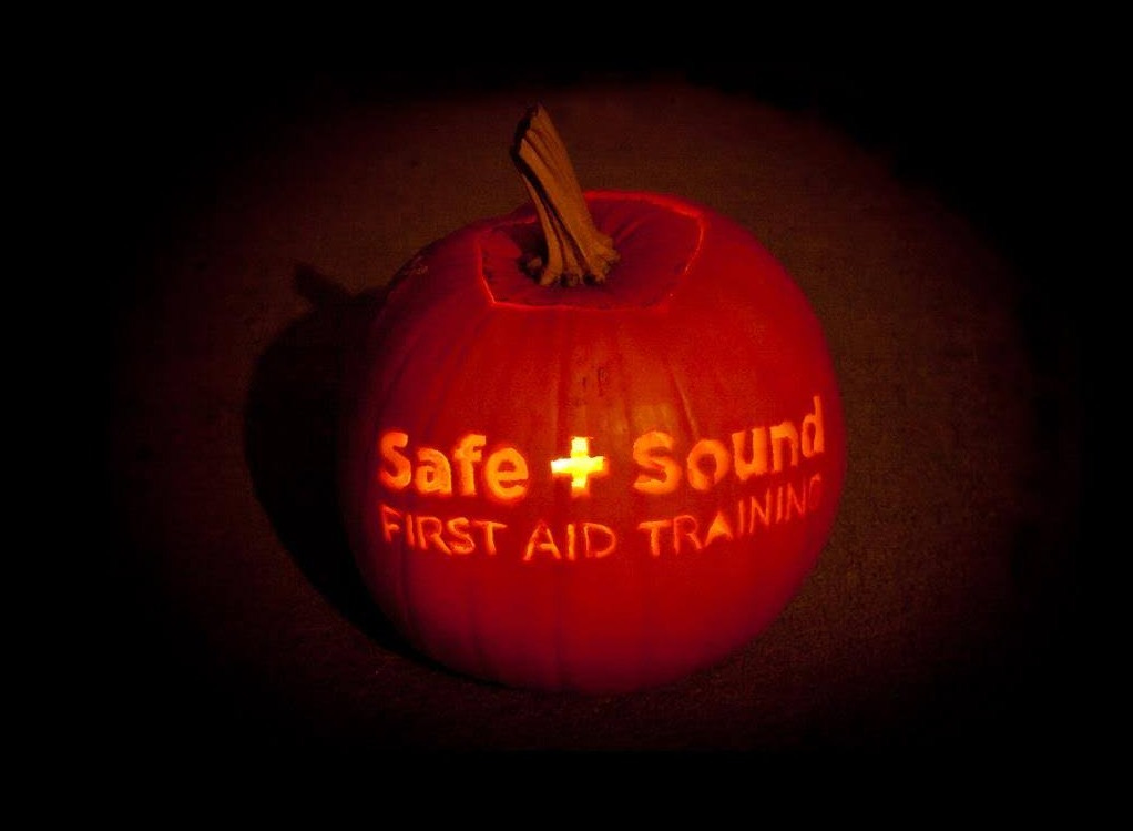 An orange pumpkin, lit from inside with the words Safe + Sound First Aid Training carved on the outside, against a dark background.