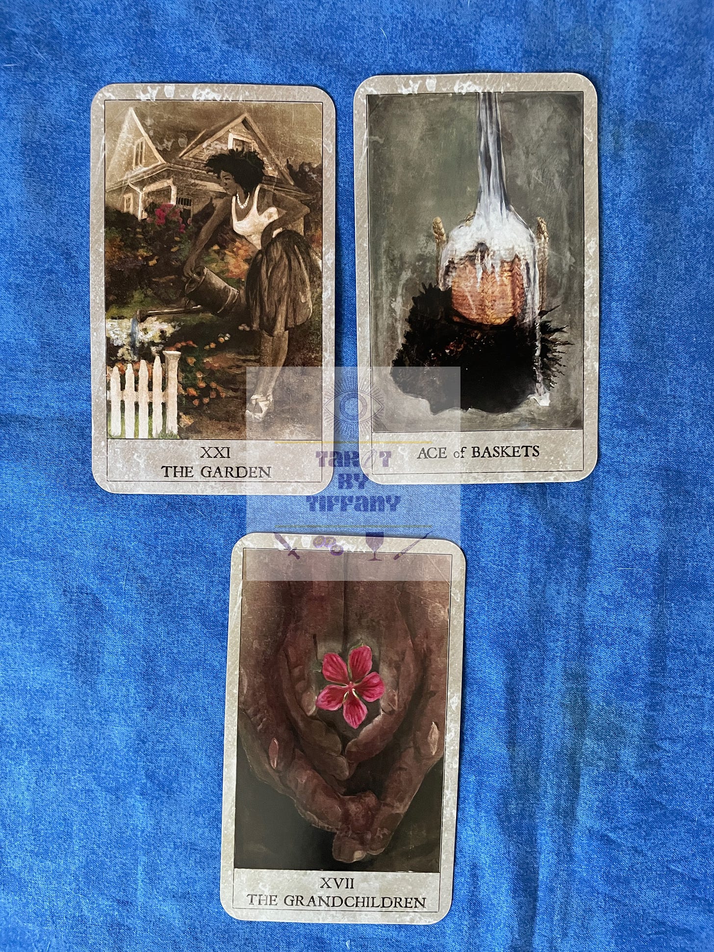 3-card reading using the Hoodoo Tarot. The cards are laid against a cobalt blue cloth background. Top two cards (L-R): XXI - The Garden and Ace of Baskets. Bottom card: XVII - The Grandchildren.