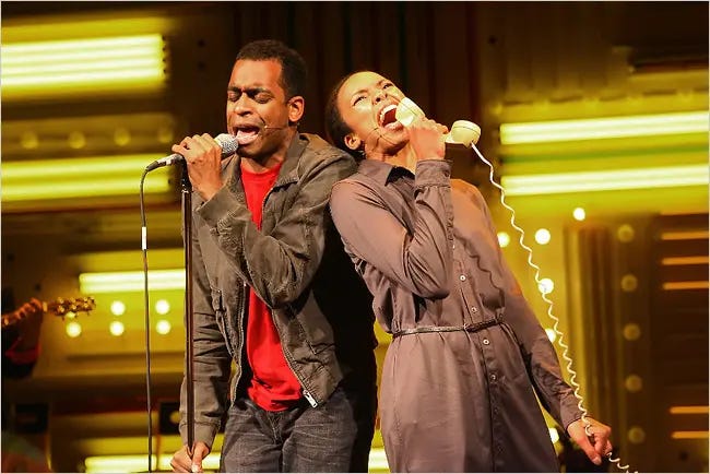 A man wearing a red shirt and a black jacket sings at a microphone, and a woman holding a corded telephone sings into the telephone next to him.