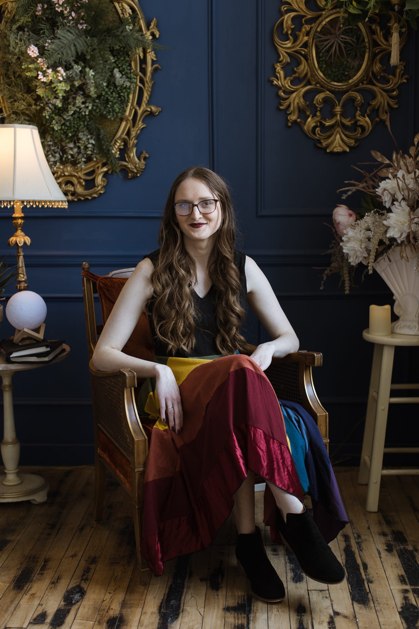 Kandi Zeller (she/her), a white woman with glasses and long reddish brown hair, sits in a burnt orange vintage chair. Kandi is wearing a rainbow dress, a dramatic red lip, and black boots. Behind her is a blue wall with two gigantic gold frames, overflowing with 3D green foliage. Next to her are two side tables. One has a stack of books and a mini moon lamp. The other has a battery-operated candle and some silk flowers.