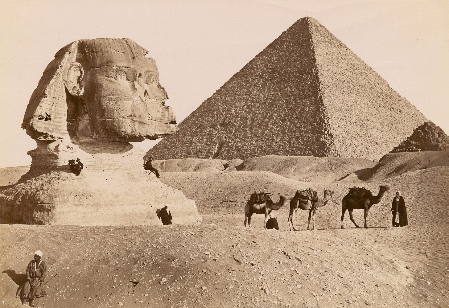 A sepia-toned photo of a pyramid and sphinx with camels and several men in front of them.