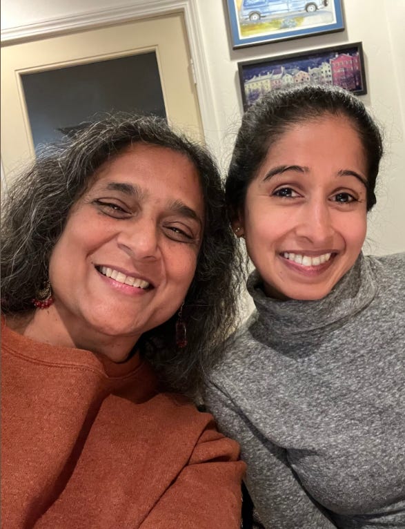 In this photo the two women, both South Asian with dark brown hair and beautiful smiles are putting their heads together and smiling at the camera.
