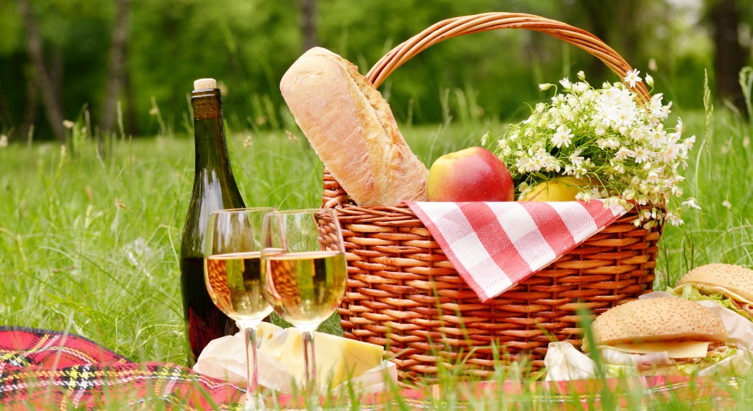 A woven picnic basket with bread, apples, and elderflower in it, with two wine glasses, sandwiches, and a checkered red blanket on the green lawn