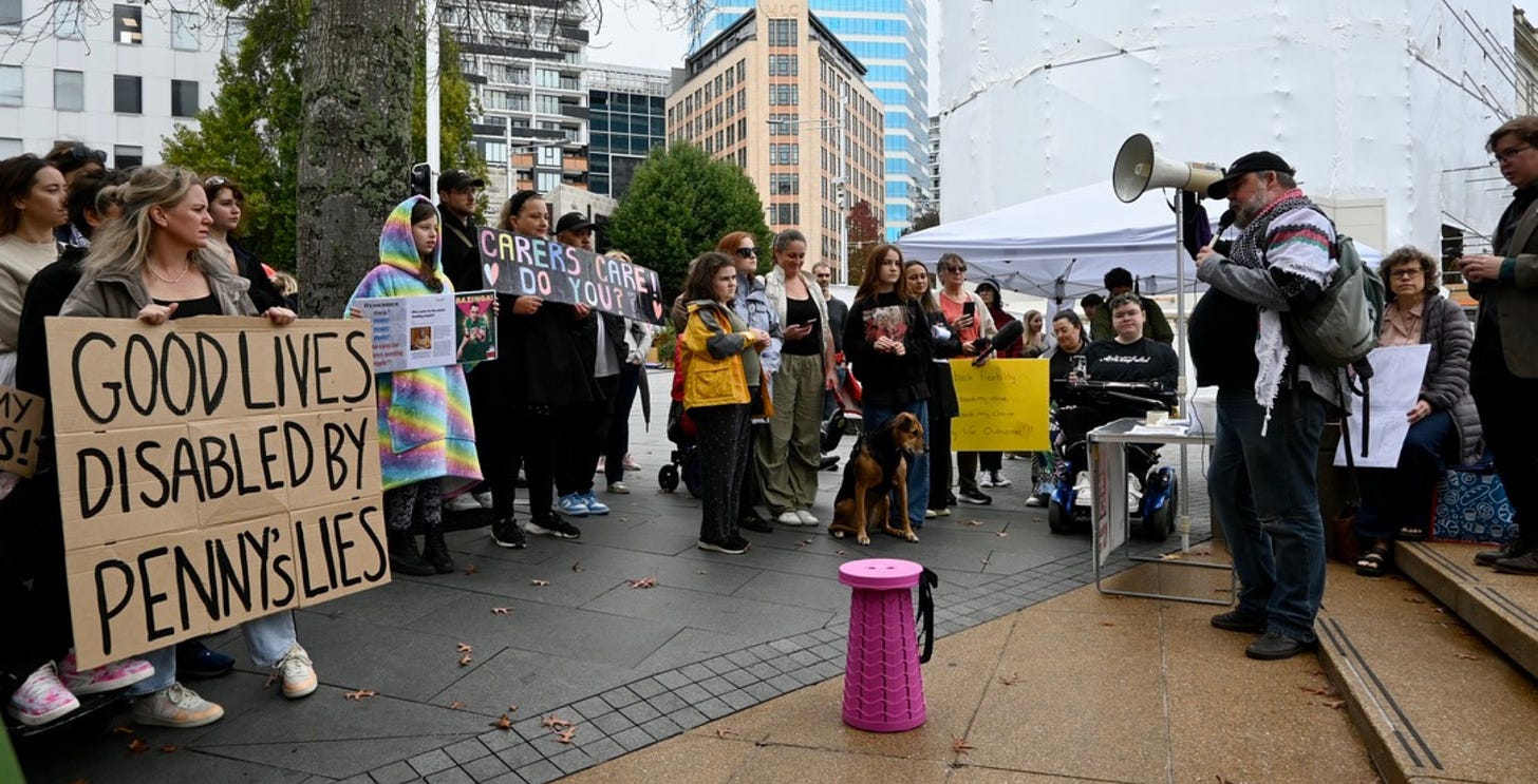 Image description: A group of disabled and nondisabled people protest, with a man speaking into a megaphone in front of the crowd.