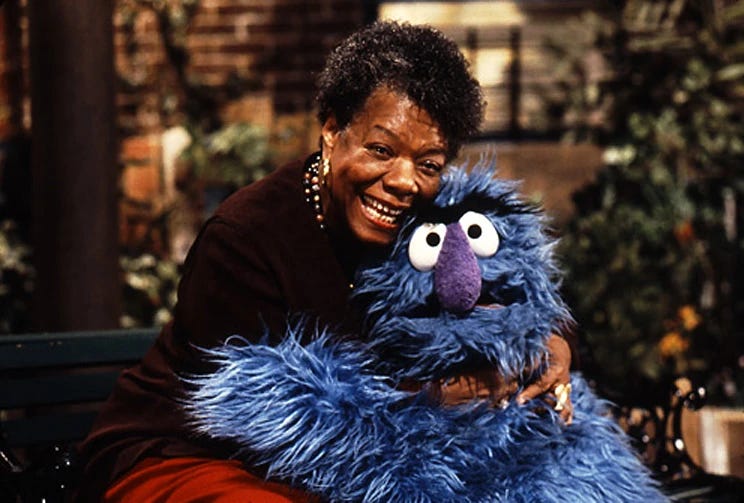 maya angelou, an older black woman wearing her hair short and a burgundy cardigan, smiles and holds a blue puppet in a hug.