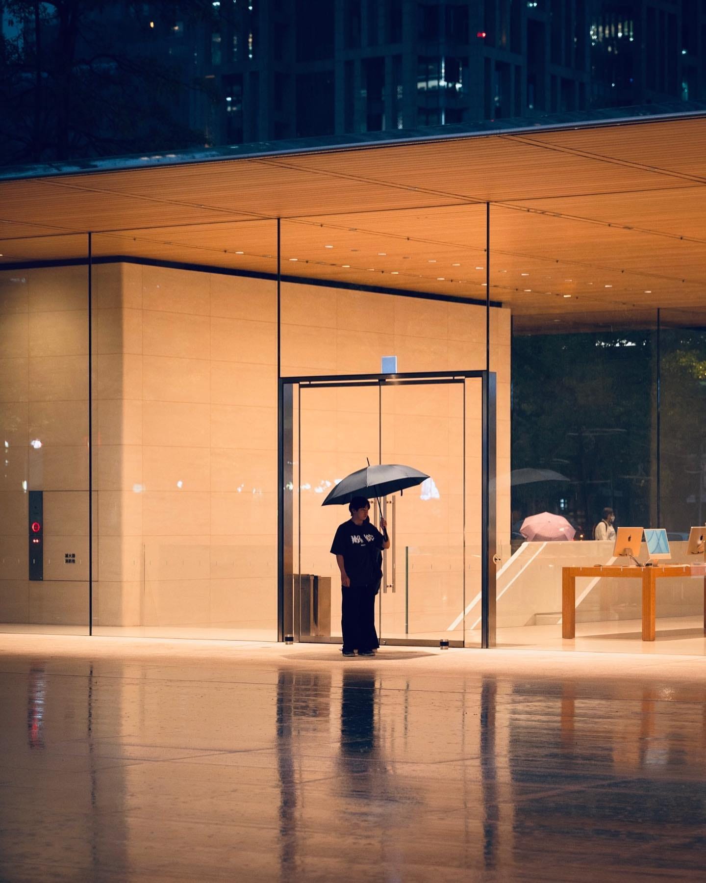 A person with an umbrella stands outside the entrance to Apple Xinyi A13 after dark.