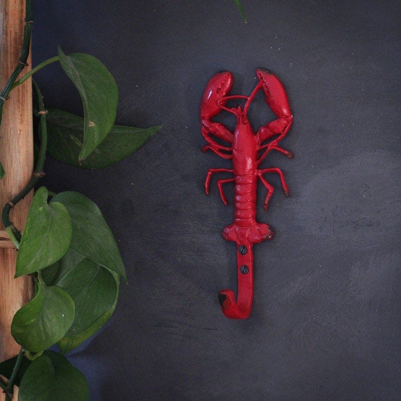 May include: Red metal lobster hook.  The lobster is detailed with claws, body segments, and eyes. The hook is attached to the lobster's body. The lobster is mounted on a dark gray background.  A green plant is partially visible in the left corner.