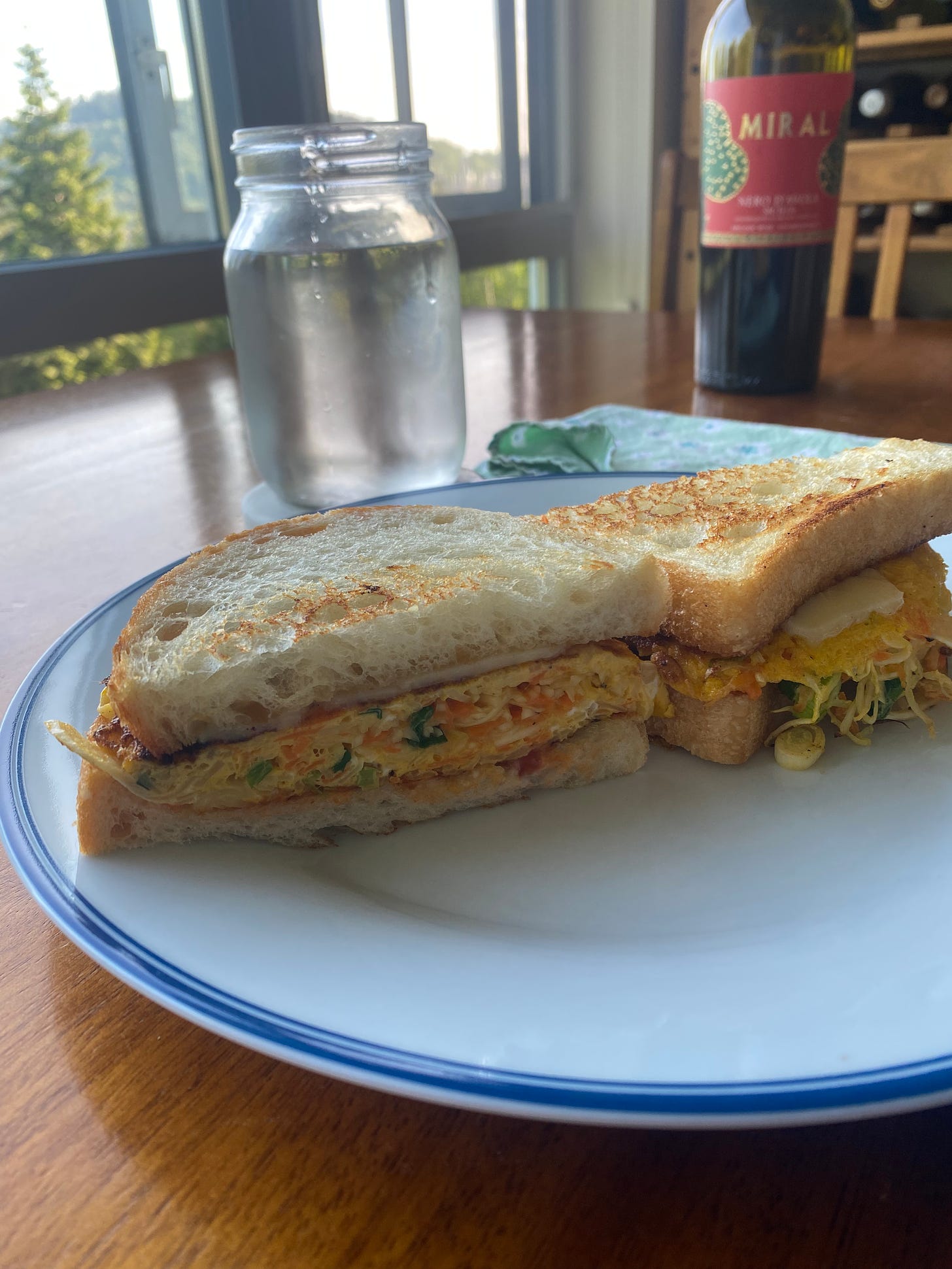 The sandwich described above: egg, carrot, cabbage, and green onion topped with cheese on toasty white bread. It's cut in half with one half facing the camera, on a blue-rimmed white plate. In the background is a bottle of wine and a glass of water.