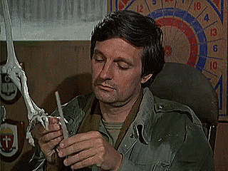 Alan Alda as Hawkeye Pierce in MASH sitting in a chair and looking up as he files the nails of a full-size plastic skeleton.