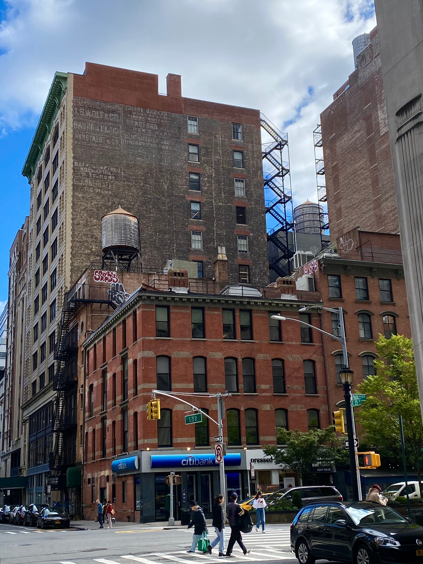 A low red brick building with a Citibank in in the store front. East of it on 25th street is a taller brick building with a water tower on its roof. The taller building east of that has faded advertisements on the brick exterior. South of the Citibank is another brick building beside a taller building. A water tower can be seen between the two tallest buildings rear fire escape stairs.