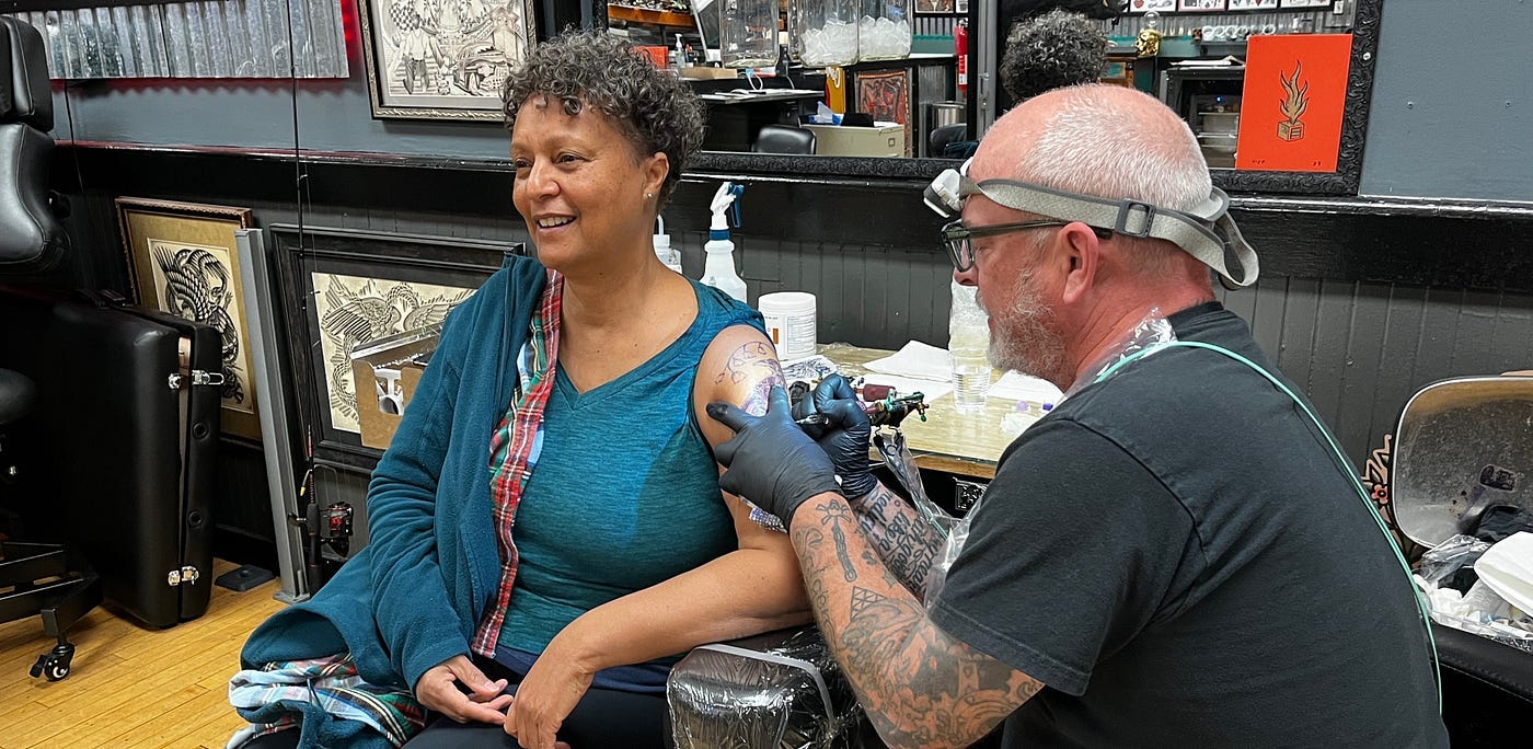 Me in the chair — smiling. Tattoo artist at work with his gun, headlamp, latex gloves