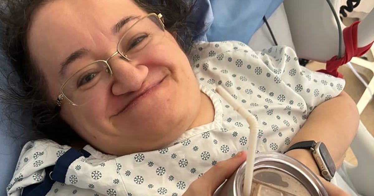 Photo of Athena smiling up from her hospital bed. She has a covered mug with a straw poking out in her hands.