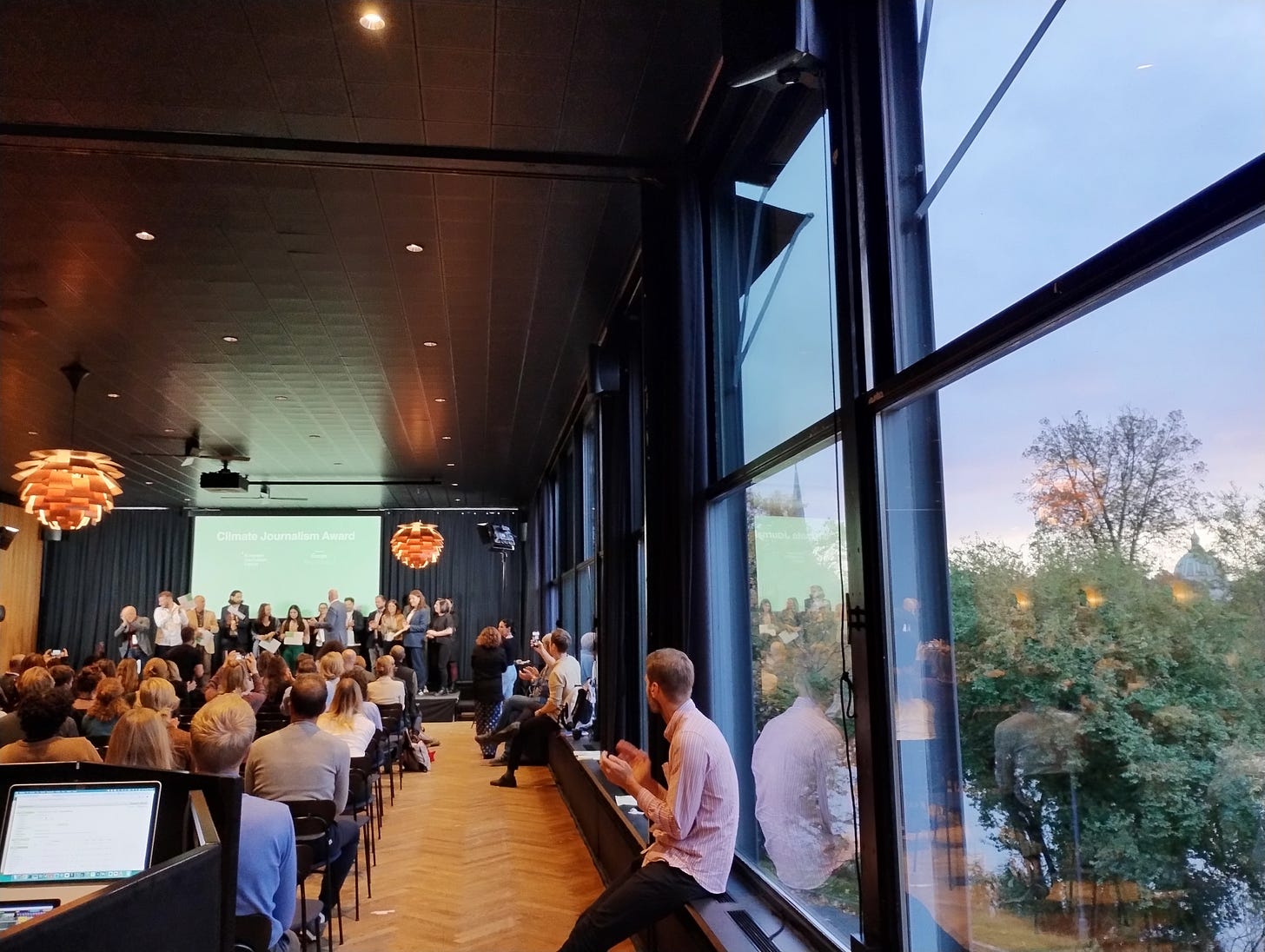 Photo of a conference venue taken from the back of the hall. Several people are accepting awards on the podium. On the right there is a large window with trees outside. The audience are applauding.