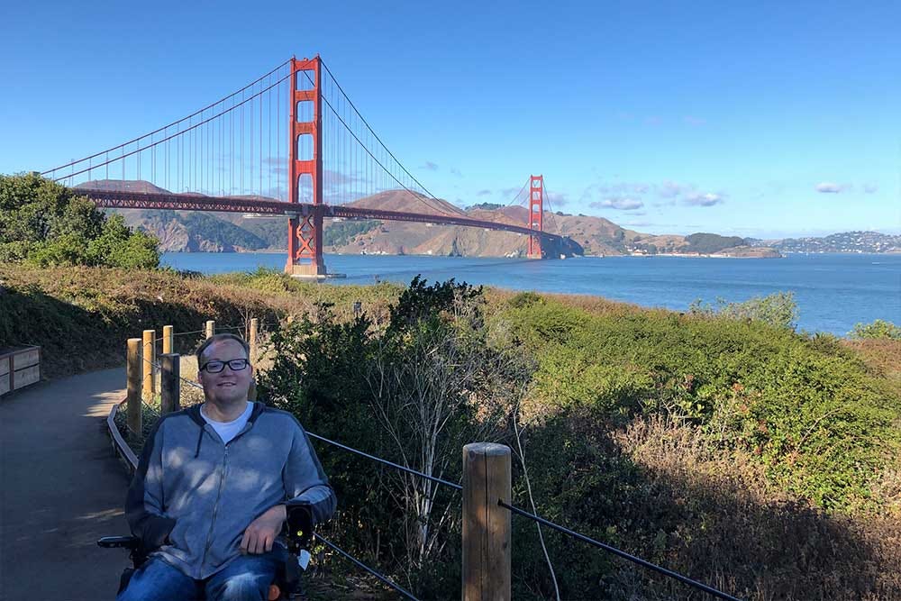 John on a paved pathway seated in his wheelchair with the Golden Gate Bridge in the background.