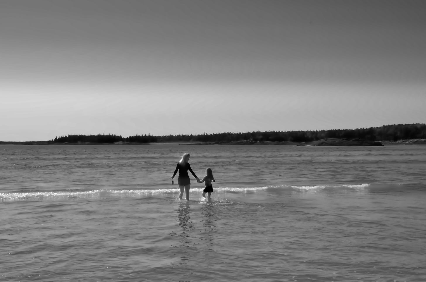 A black and white image of a mom and daughter in the distance splashing in shallow waves on the coast.